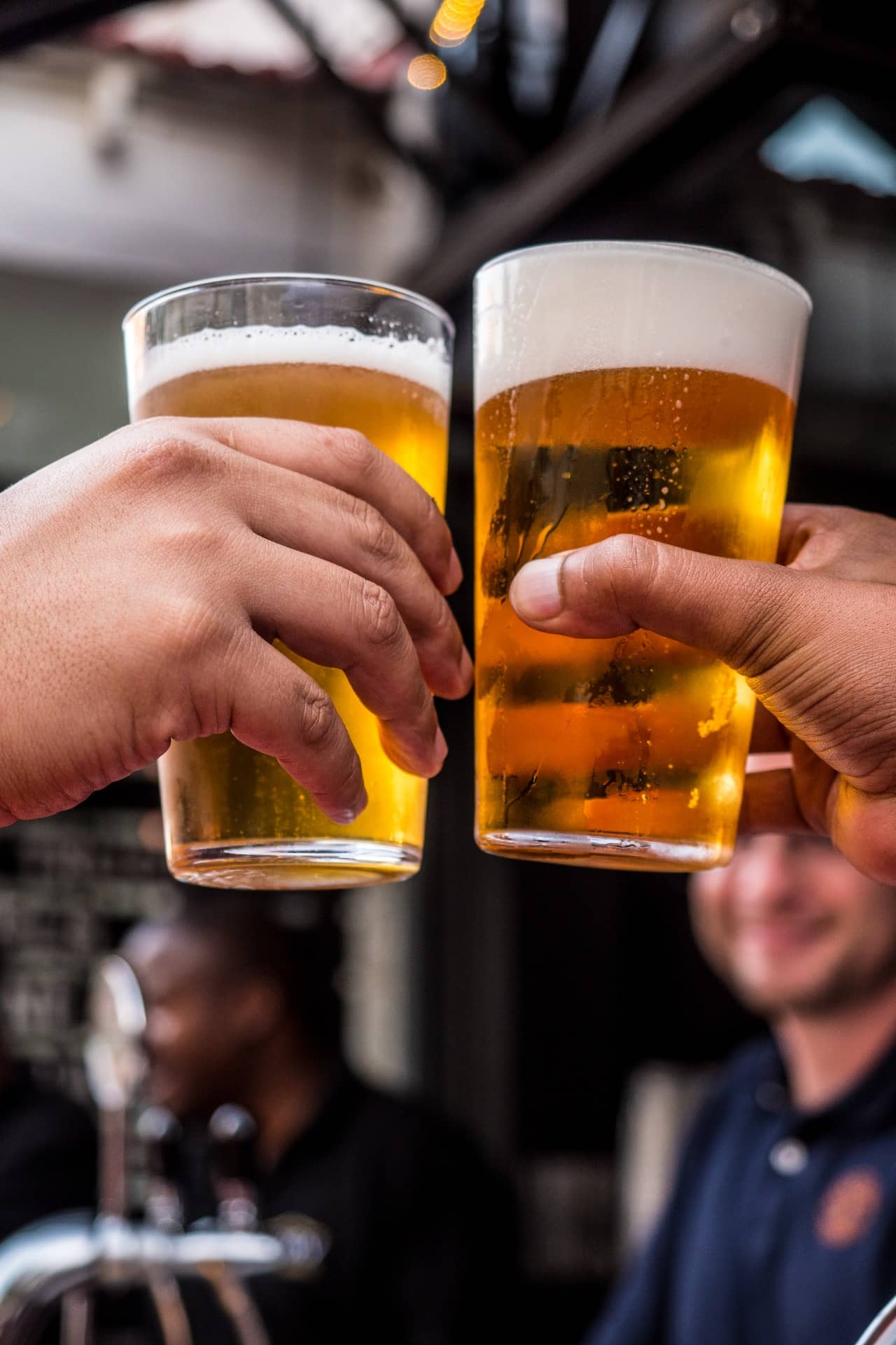 vertical photo showing two hands holding beer glasses in 'cheers' motion with out of focus people in the backgound