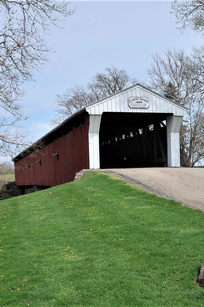vertical photo of the eldean covered bridge showing the road to the bridge and a wide grass bank. There are trees in the background