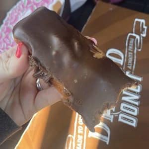 square photo showing a hand holding a bitten-into chocolate iced gluten-free finger donut from gluten-free from the dipped
