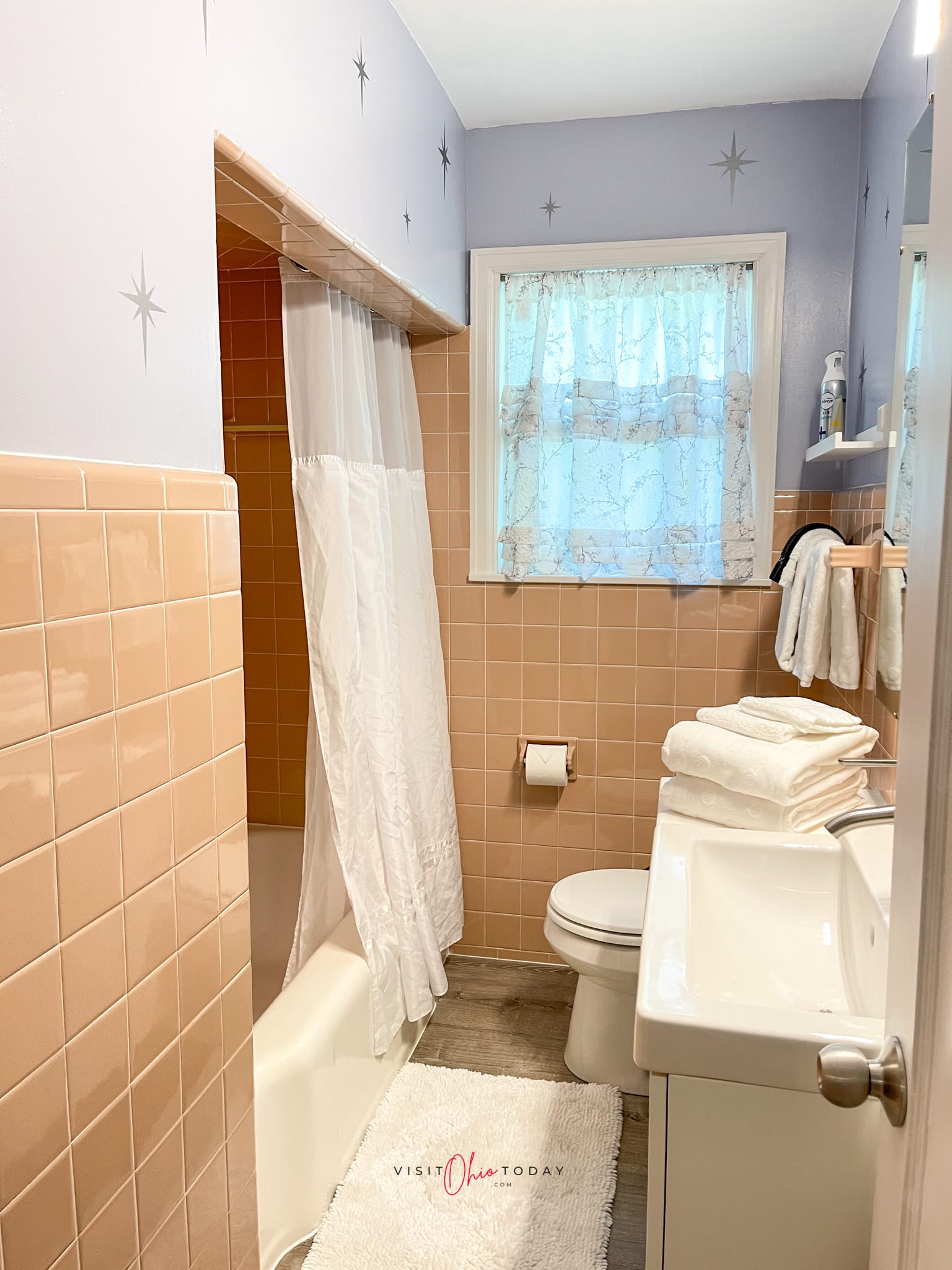 light pink tiled bathroom on tub and floors, light purple paint on walls with silver stars (all on left) on the right is a white skinny sink, white towels and toilet
