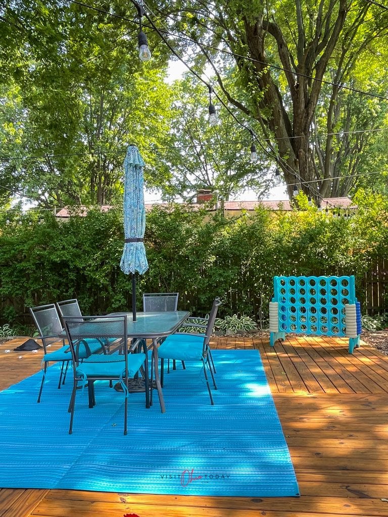 backyard with a wooden deck, blue rug, 6 person glass table with umbrella and a giant connect 4 game