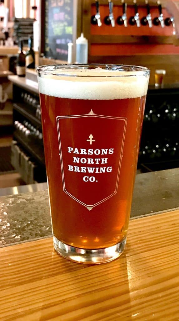 vertical photo showing a large glass of beer with Parsons North Brewing Co, on a bar with beer taps in the background