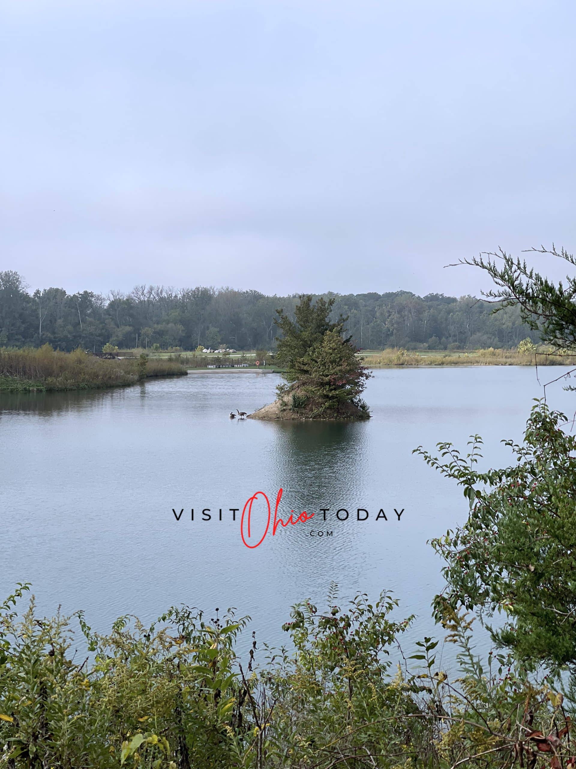 vertical photo showing a lake at prairie oaks metro park with a small island with foliage on it, and foliage in the foreground Photo credit: Cindy Gordon of VisitOhioToday.com