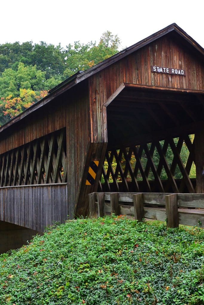 vertical photo of State Road Covered Bridge with a grassy hillock in the foreground and trees in the background