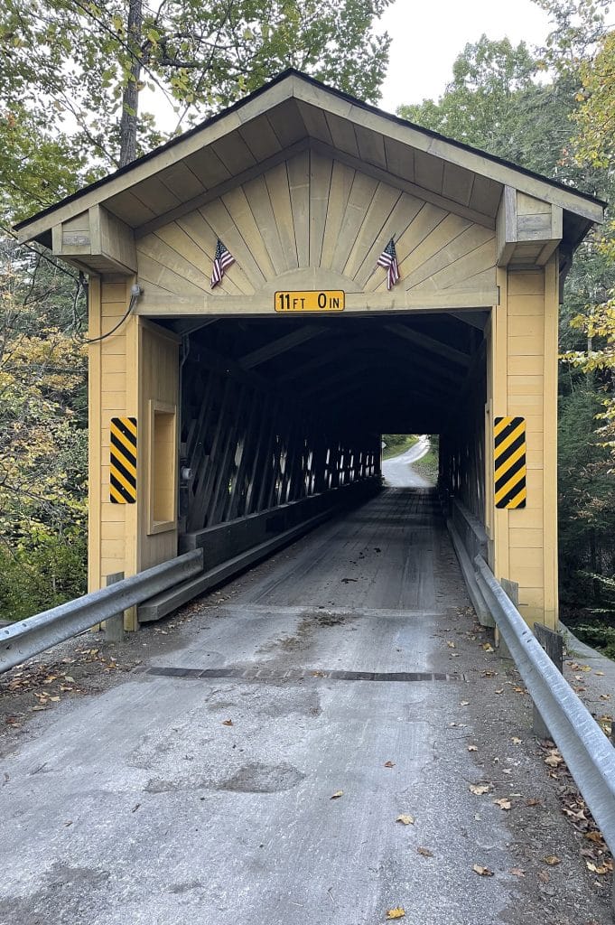 vertical photo that is a shot straight through windsor mills covered bridge, with a road in the foreground and trees in the background