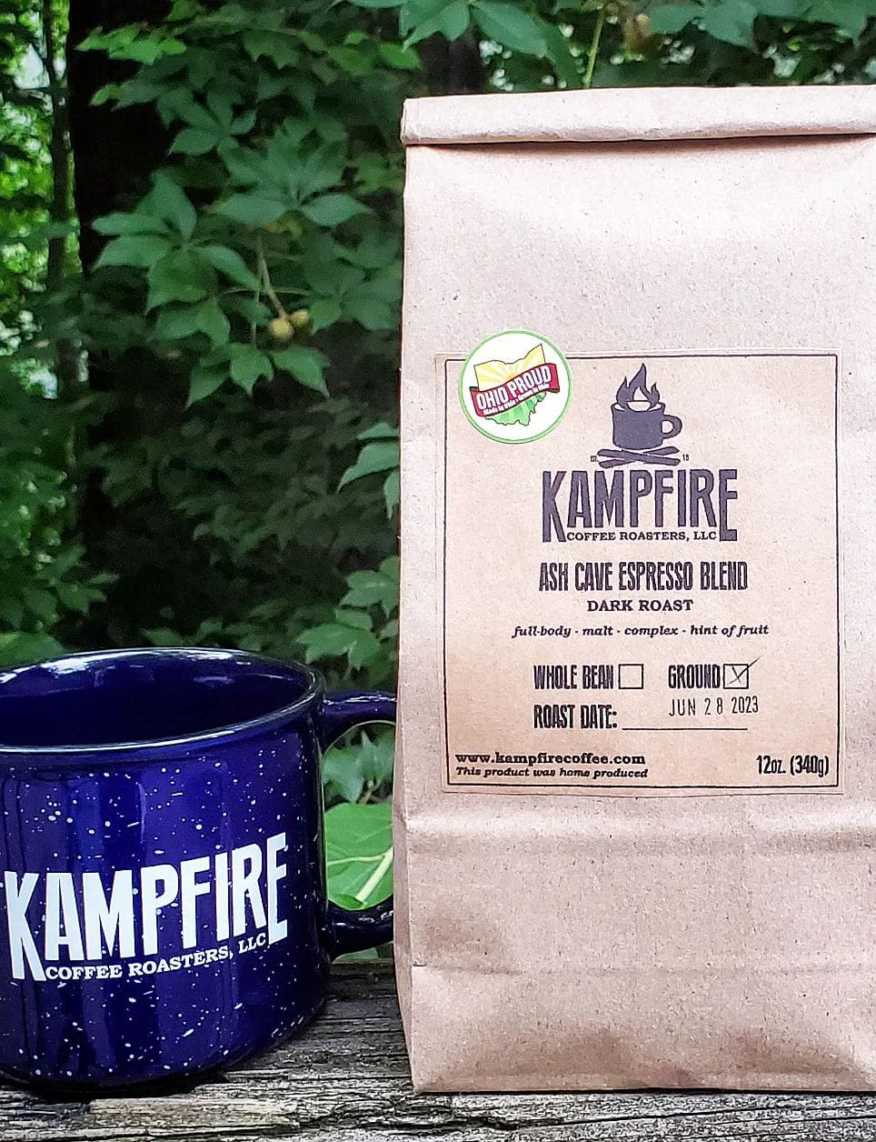 horizontal photo of a bag of Kampfire Ash Cave Espresso Blend coffe, with a blue Kampfire coffee mug beside it, on an outdoor wooden table. A large tree with lots of green foliage is in the background. Image courtesy of Kampfire Coffee Roasters
