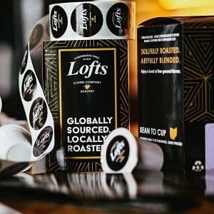 square photo of two bags of lofts coffee company coffee, with a coffee pod and some lofts round stickers on a roll. Image courtesy of Lofts Coffee Company and Roastery