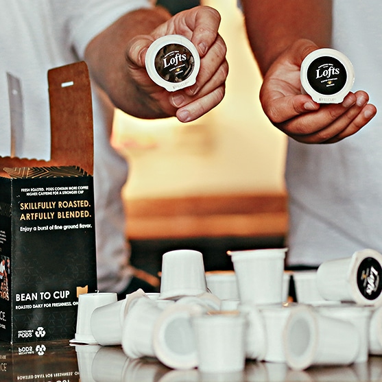 square photo of two hands each holding a lofts coffee company coffee pod with more coffee pods on a wooden surface in the foreground. Image courtesy of Lofts Coffee Company and Roastery