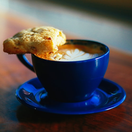 square photo of a cup of coffee in blue cup and saucer with a cookie resting on the edge and the handle of the cup. Image courtesy of Lofts Coffee Company and Roastery