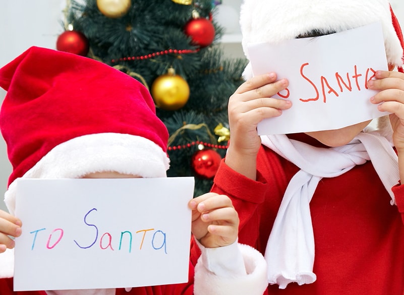 horizontal photo of two children holding letters to Santa in front of their faces with a christmas tree in the background