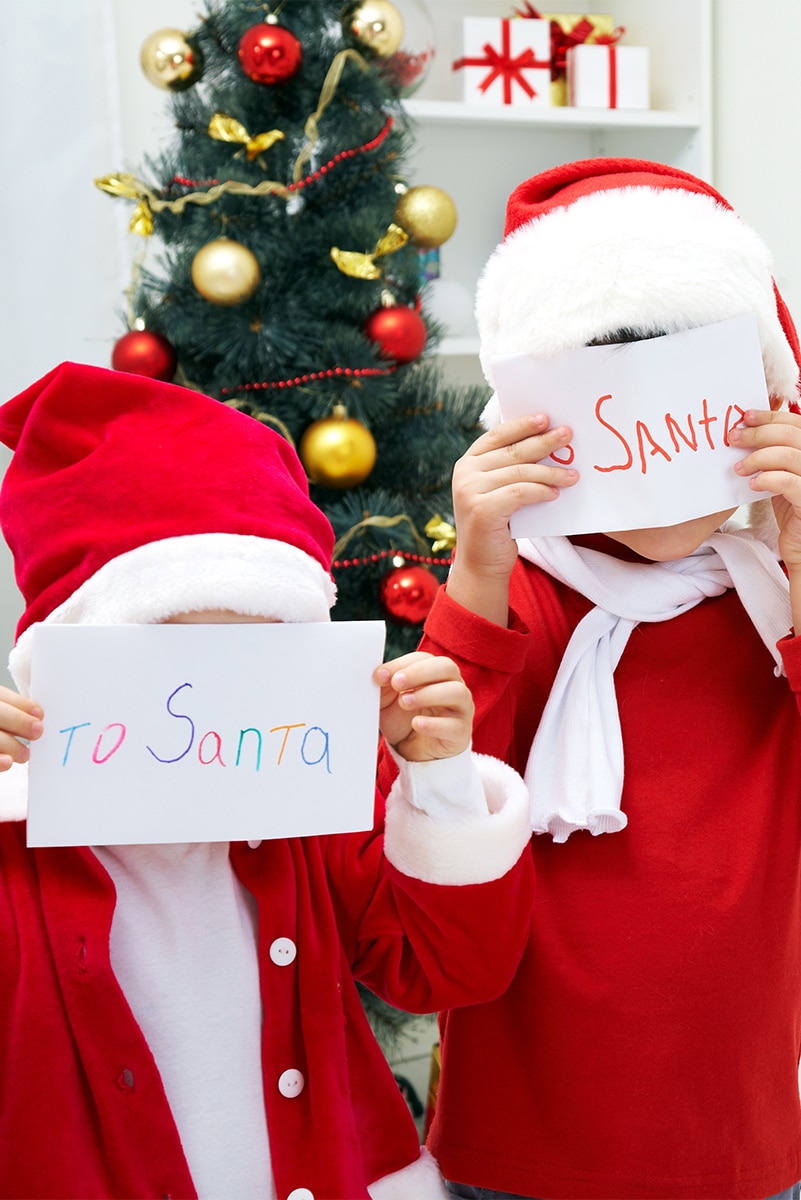 two children in red and white clothes with santa hats standing in front of a green christmas tree with white, red and gold bulbs. Holding letters addressed to santa in front of their faces