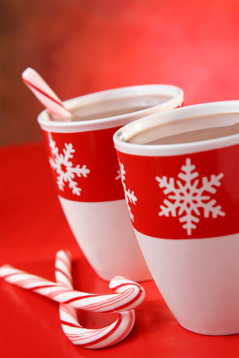 vertical photo of two mugs with hot chocolate. One mug has a candy cane in it and there are 2 candy canes beside the mugs. All on a red background