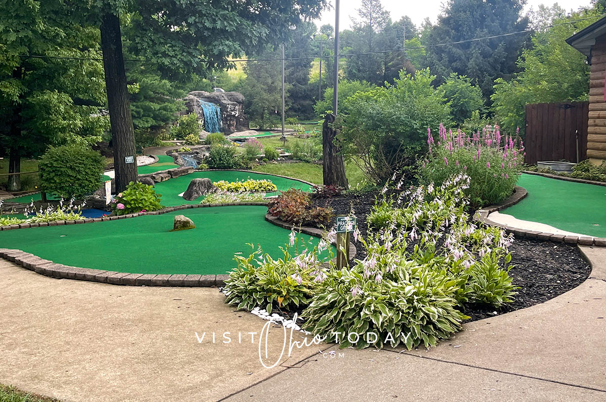 horizontal photo of an area of Adventure Golf at Rempel's Grove with the footpath in the foreground and plants, trees and grass surrounding the area