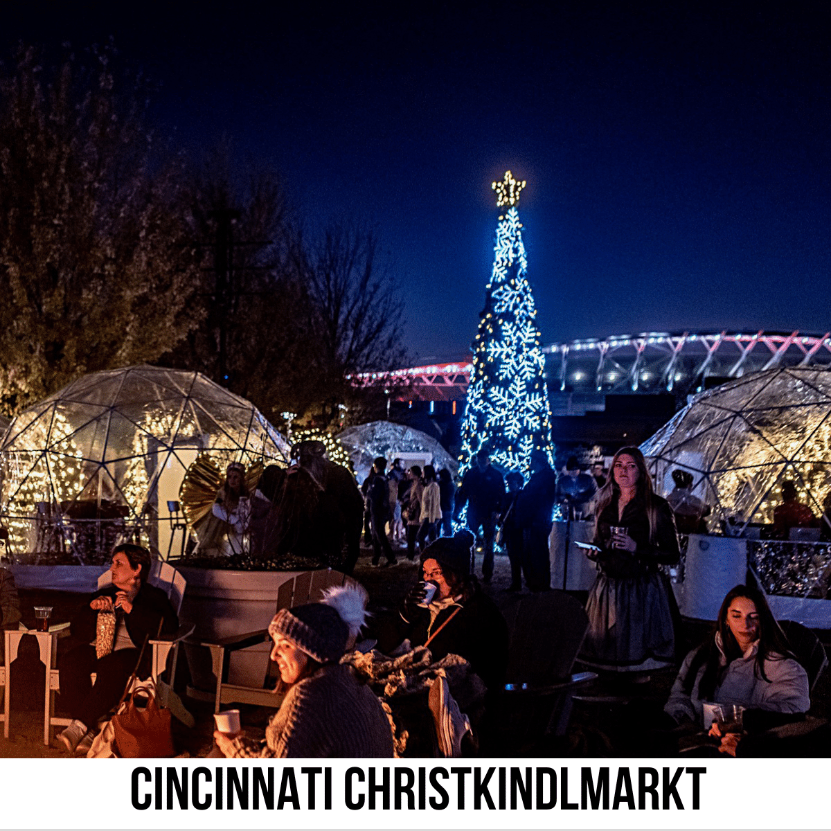 square image with a photo of the Cincinnati Christkindlmarkt with lit igloos and the lit Christmas tree and people sitting around an open fire in the foreground. A white strip across the bottom has the text Cincinnati Christkindlmarkt. Image courtesy of Cincinnati Christkindlmarkt