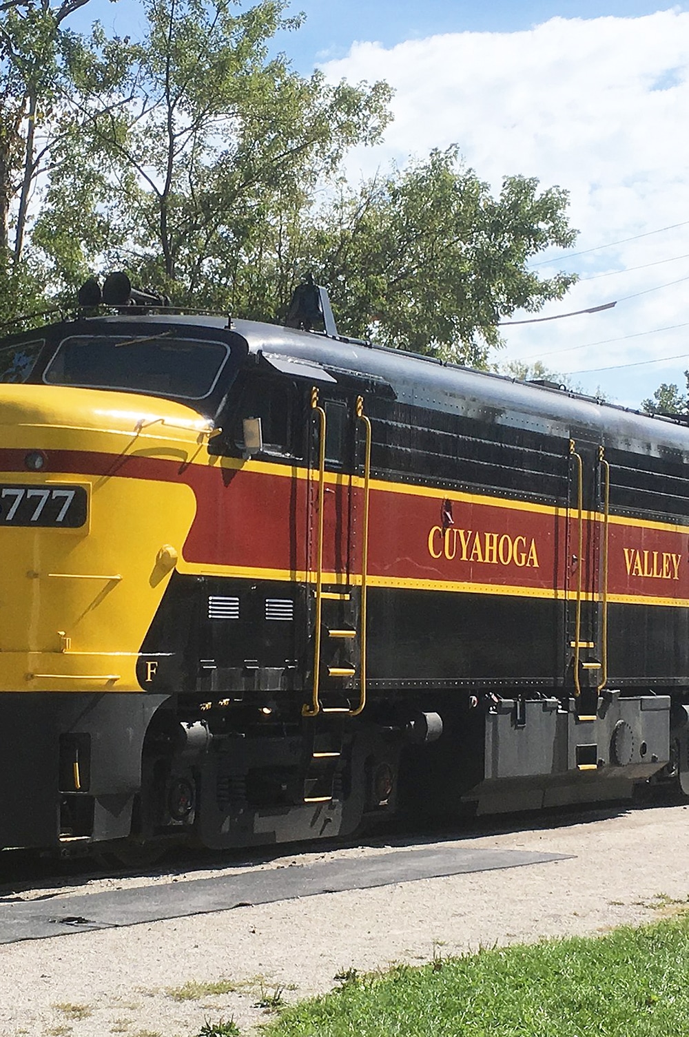 vertical photo of a train with Cuyahoga Valley on the side, and the number 6777 on the front. trees behind and grass in front. Image via Wikimedia Commons