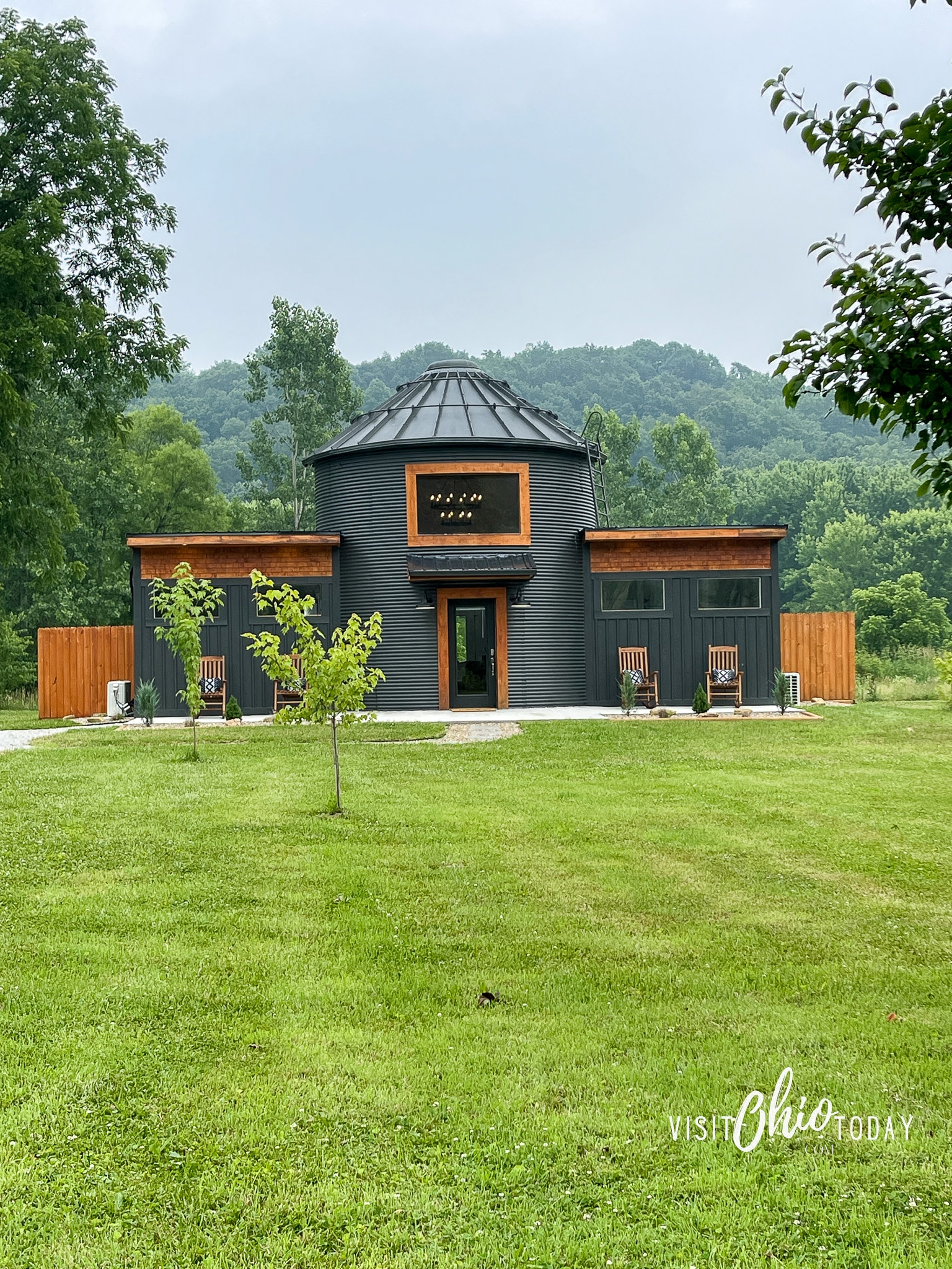 pictured is a black silo made into a tiny home. The black silo and buildings on each side are set back off the road and are in hocking hills