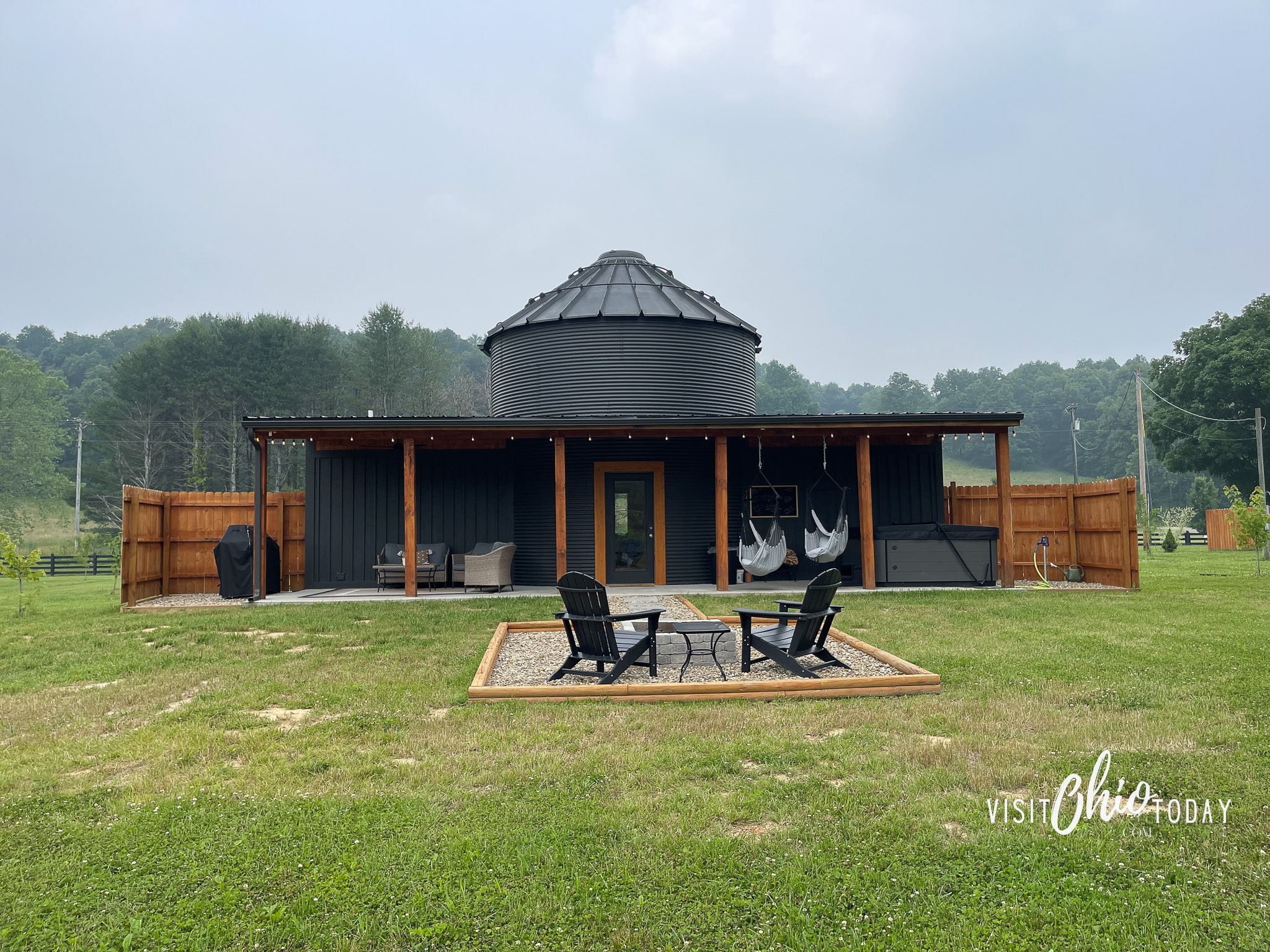 the back of the silo cottage in hocking hills. It is black with a covered patio that has two hanging chairs, a large hot tub and seating for 4 people. There is a grill and also a fire pit with two chairs.
