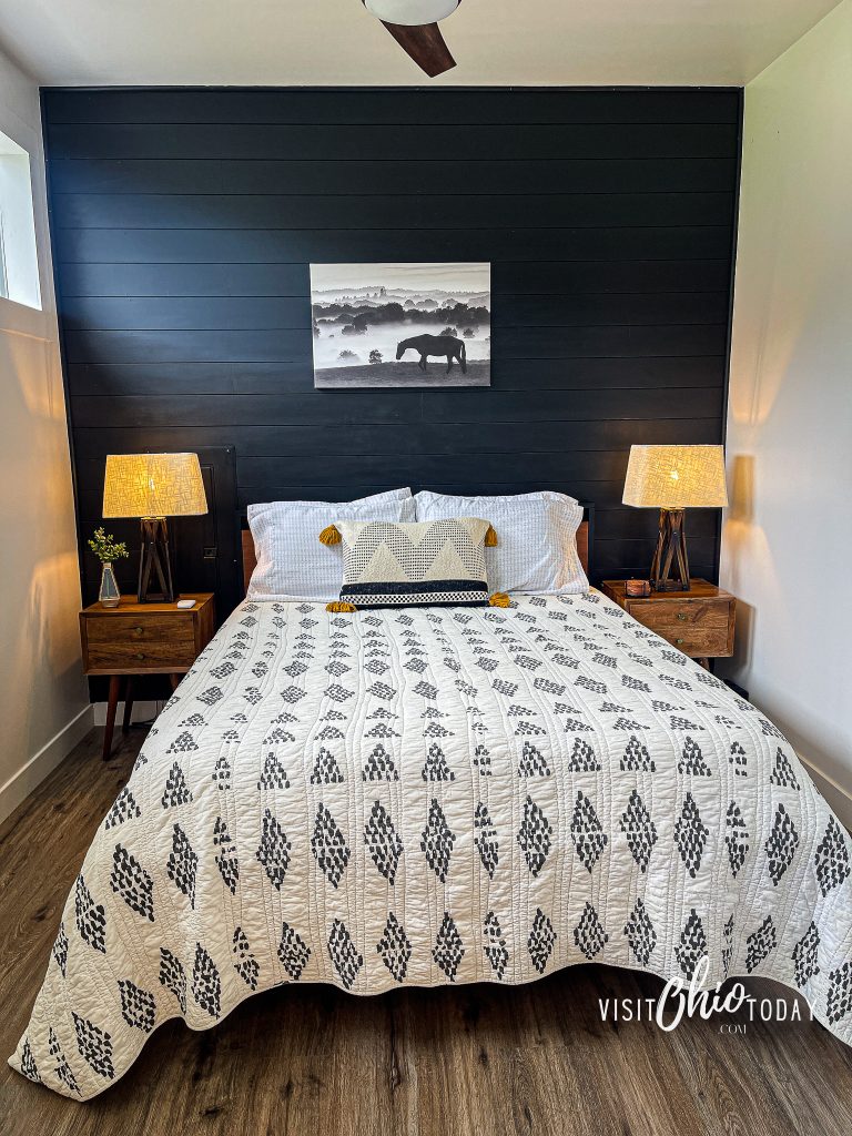 pictured is the 1 bedroom of the silo cottage hocking hills, there is a black panel wall with a queen bed and tables with lamps on each side. The bed has a black and white spread on it