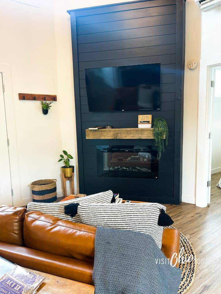pictured is the living room at the silo cottage hocking hills, you can see a small brown leather couch with a gray blanket on it, a black paneled wall with a gas fireplace