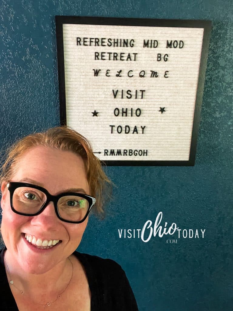 Cindy from visit ohio today with black glasses and a black shirt standing in front of the sign that reads refreshing mid mod retreat BG welcome visit ohio today