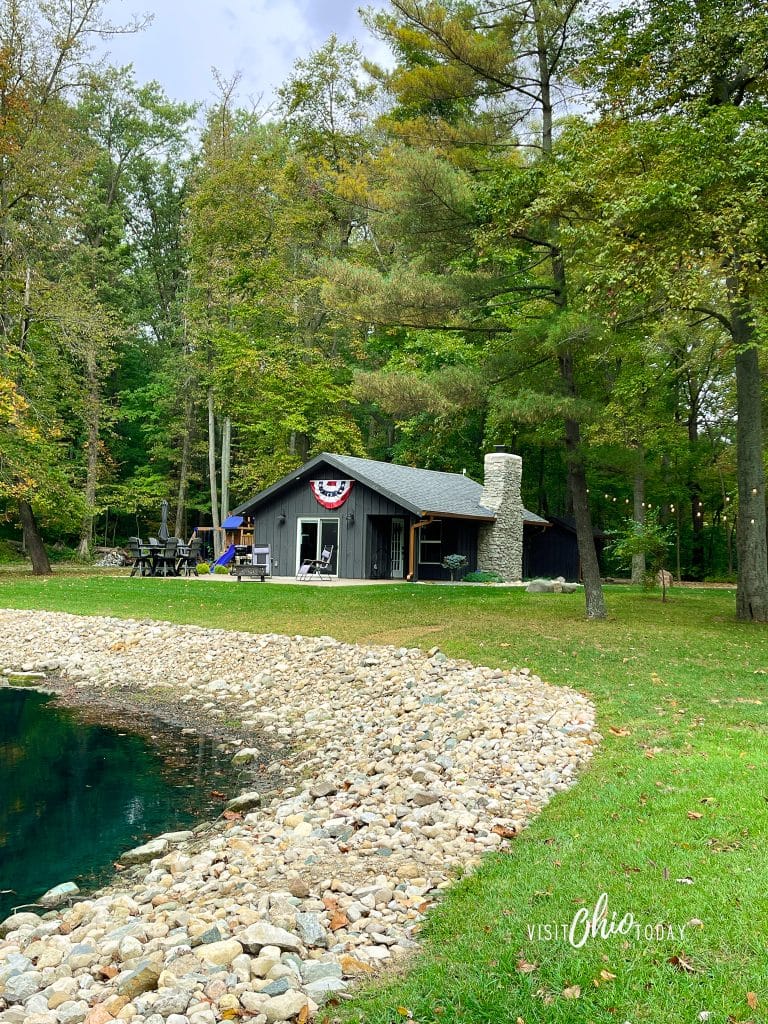 photo of a dark colored cabin in the woods with a strong chimmey, you can see the edge of a blue colored pond photo credit: Cindy Gordon of VisitOhioToday.com