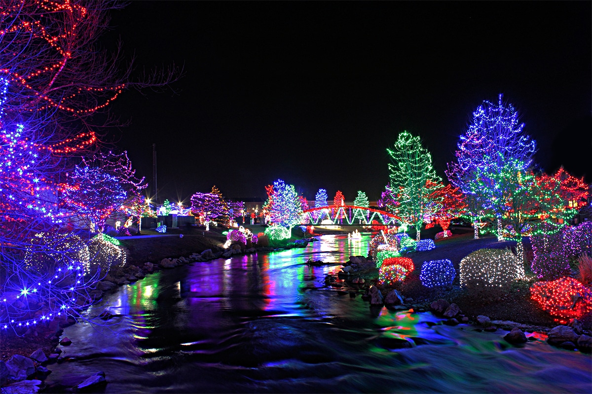 horizontal photo of drive through Christmas lights with a river in the foreground and a lit-up bridge in the background. The river banks are also decorated with lights