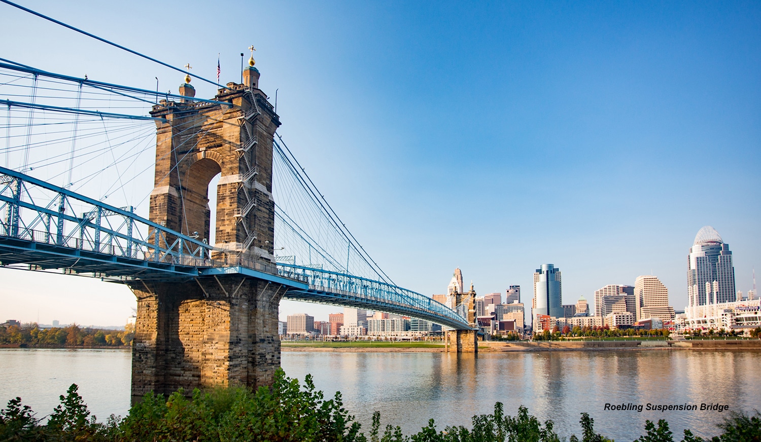 horizontal photo of the Roebling Suspension Bridge from the Kentucky side of the Ohio River, with the Ohio Cincinnati skyline in the background