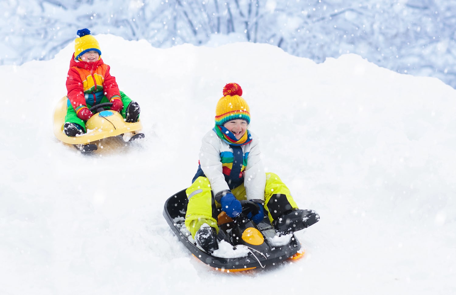 horizontal photo of two young boys in very colorful clothing, enjoying sledding down a hill