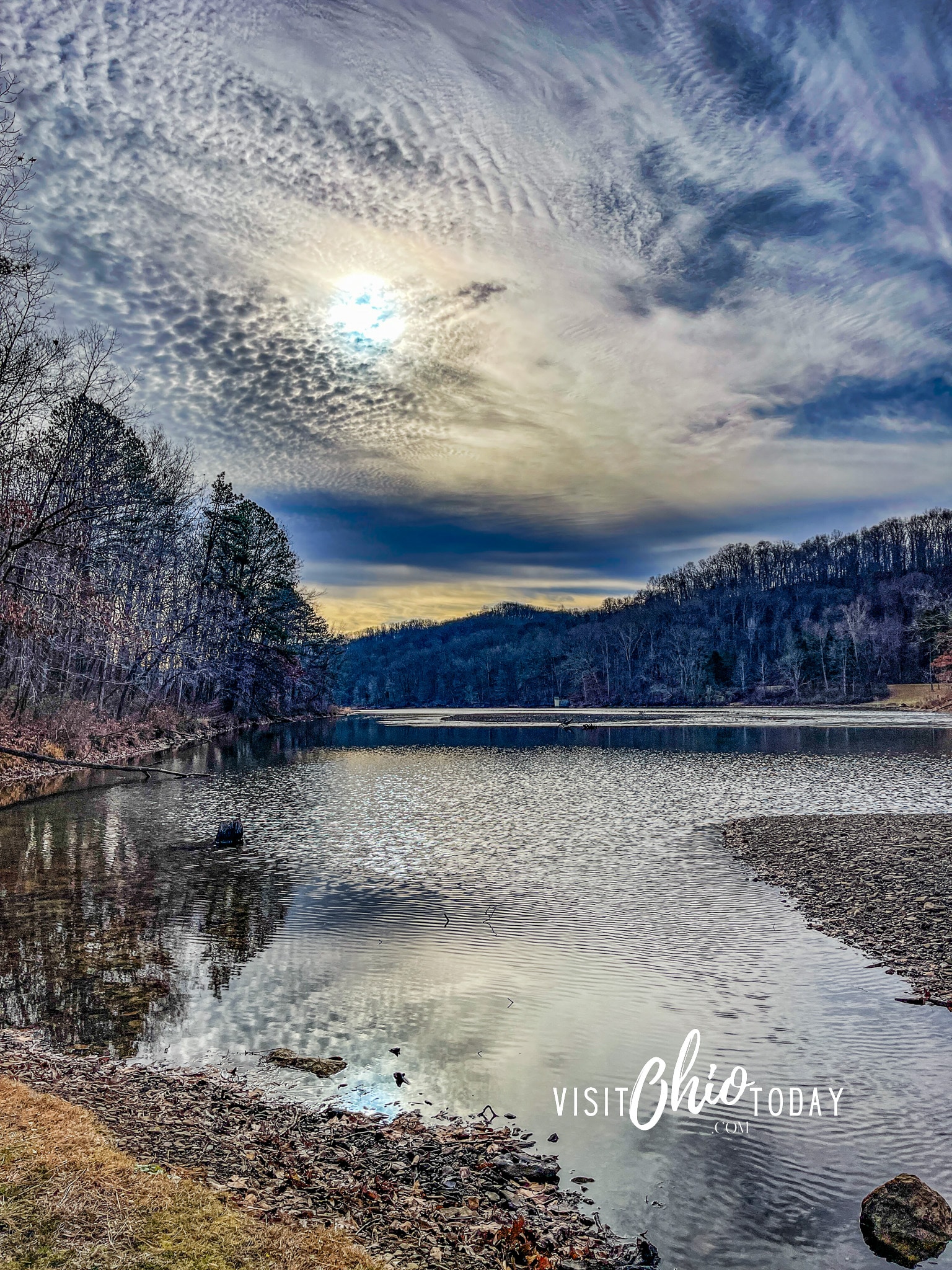 vertical photo of a lake at Shawnee State Park with trees in the background and a cloudy but blue sky. Photo credit: Cindy Gordon of VisitOhioToday.com