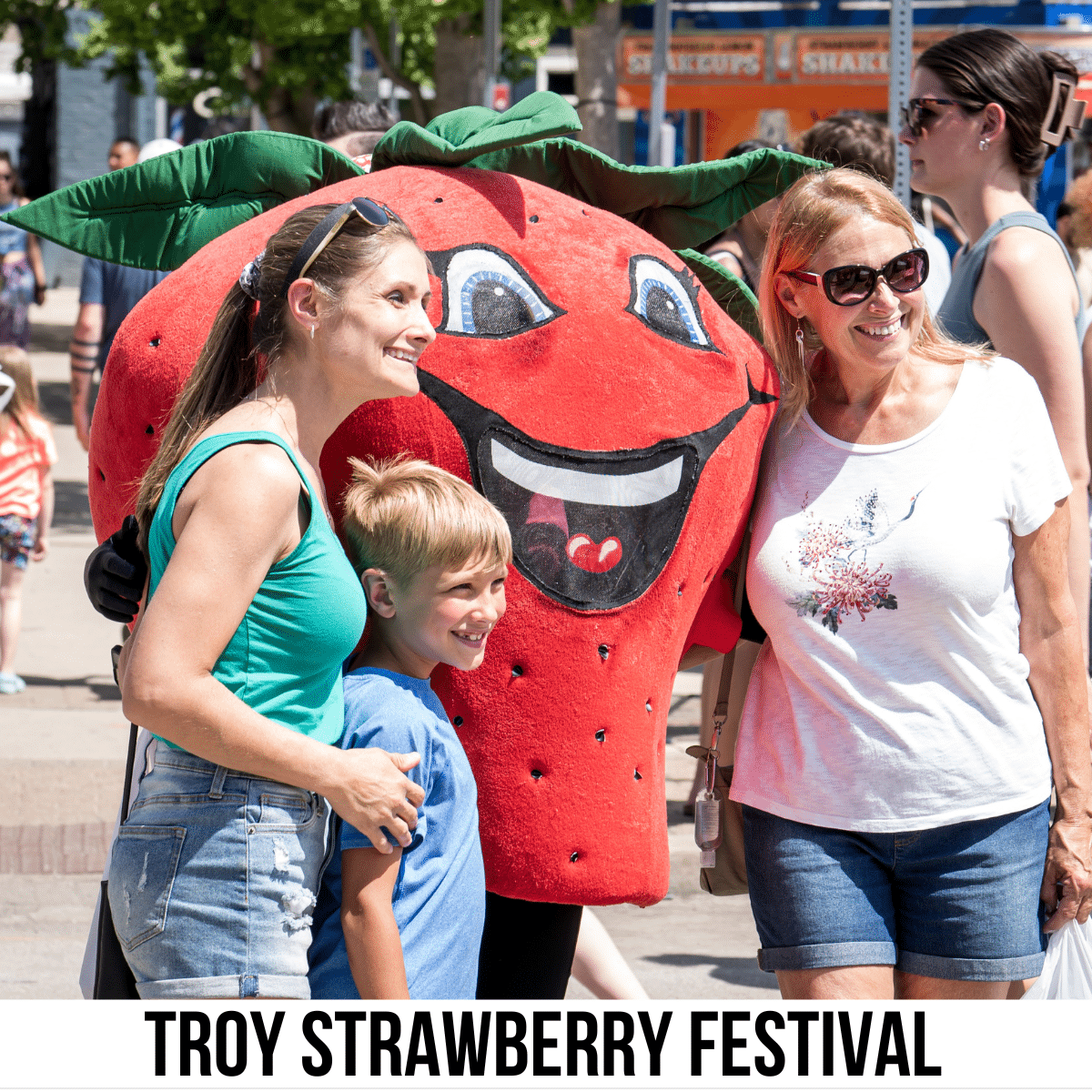 A square image of a photo of two women and a child posing for a picture with a strawberry mascot. A white strip across the bottom has text Troy Strawberry Festival.