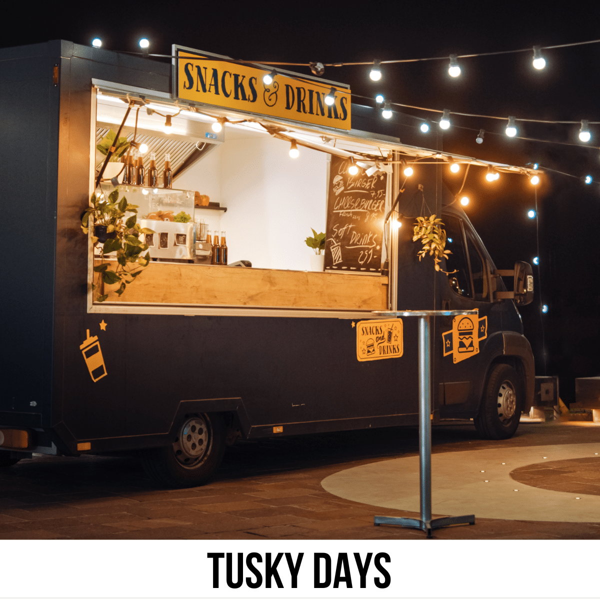 A square image of a photo of a food truck with decorative lights around it, at night. A sign above the truck window says Snacks & Drinks. A white strip across the bottom has text Tusky Days.