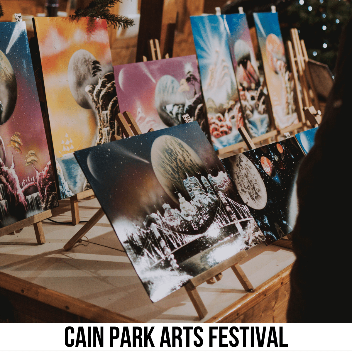 A square image of a photo of several paintings on canvas, set up on easels, on a tabletop. A white strip across the bottom has text Cain Park Arts Festival.