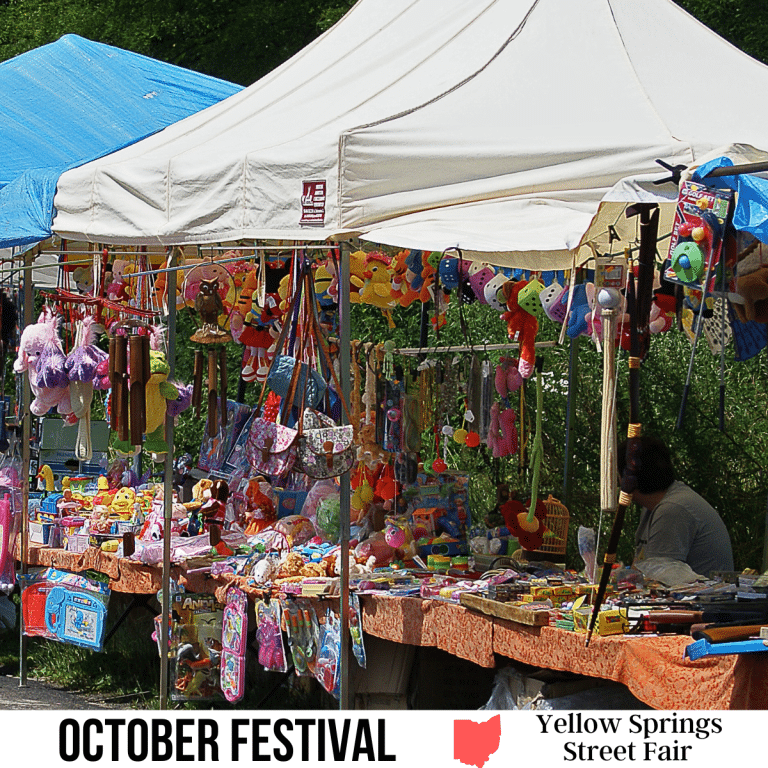 A square image of photo a vendor's booth set up at a street fair. Lots of colorful merchandise is hanging up and lying on tables under a white tent. A white strip across the bottom has text October Festival Yellow Springs Street Fair