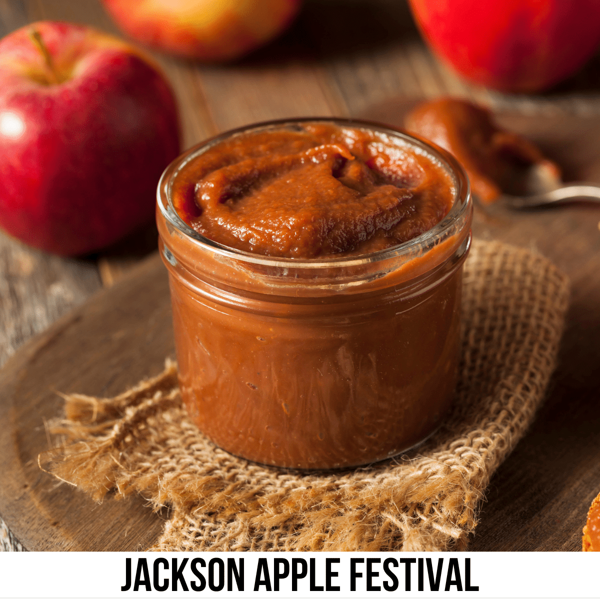 A square image of a photo of a glass jar of apple butter sitting on a piece of burlap, with apples sitting on the table in the background. A white strip across the bottom has text Jackson Apple Festival.