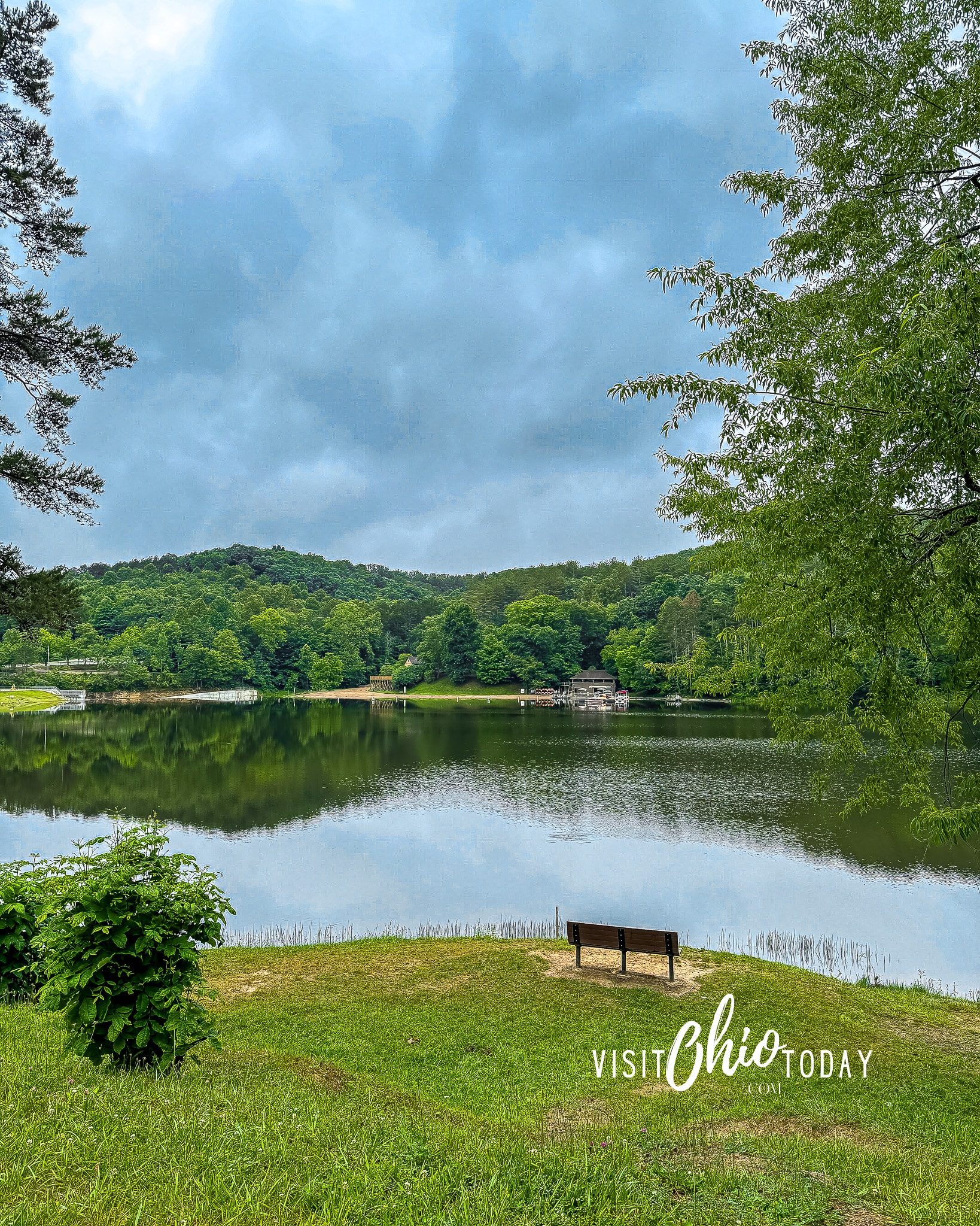 vertical photo of the lake at Lake Hope State Park, with a grass area in the foreground with a bench and trees to the sides and in the background. Photo credit: Cindy Gordon of VisitOhioToday.com