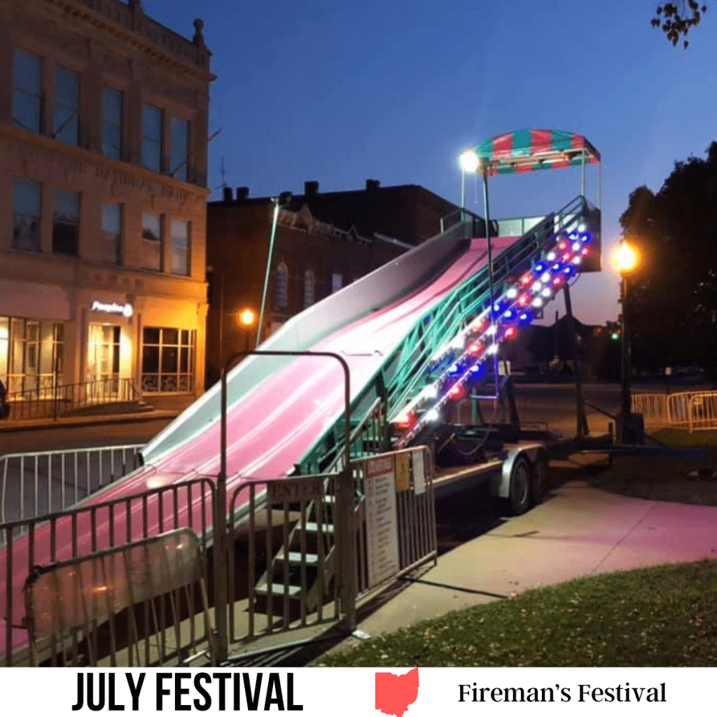 square image with a photo of a lit slide at a fairground. A white strip across the bottom has the text July Festival. Fireman's Festival