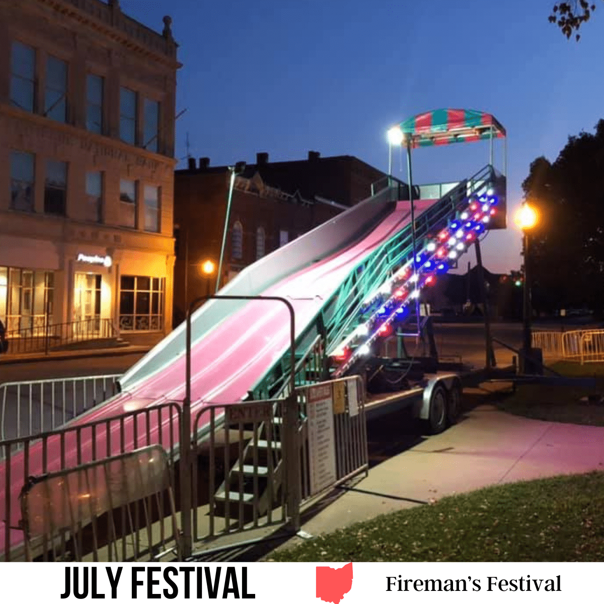 square image with a photo of a lit slide at a fairground. A white strip across the bottom has the text July Festival. Fireman's Festival