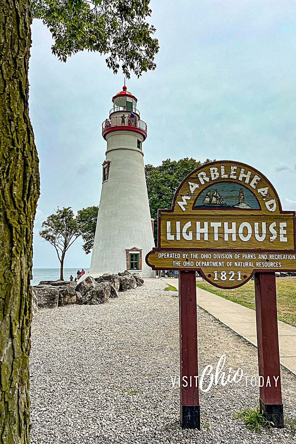 vertical photo of Marblehead Lighthouse in the distance and the entry sign in the foreground. Photo credit: Cindy Gordon of VisitOhioToday.com